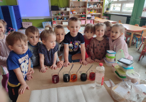 Grupka uśmiechniętych dzieci, stoi przy stoliku. Na stoliku jajka ,oraz szklanki z kolorowym płynem do farbowania jajek.