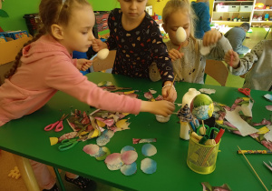 Przy stoliku dzieci, trzymają w ręku jajko ze styropianu. Na stoliku klej i kolorowe ozdoby.