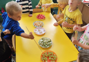 Dzieci stojące przy stole. Własnoręcznie przygotowujące szaszłyki owocowe.