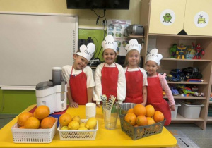 Dzieci z grupy biedronek pozujące do zdjęcia w fartuszkach,przy stole z owocami do zrobienia soku.