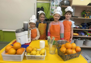 Dzieci z grupy biedronek pozujące do zdjęcia w fartuszkach,przy stole z owocami do zrobienia soku.