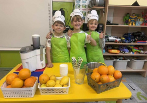 Dzieci z grupy biedronek pozujące do zdjęcia w fartuszkach,przy stole z owocami do zrobienia soku.