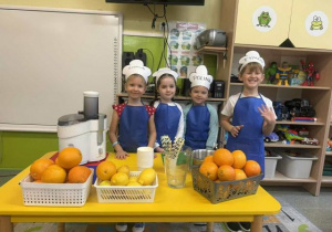 Dzieci z grupy biedronek pozujące do zdjęcia w fartuszkach,przy stole z owocami do zrobienia soku.