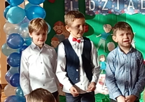 Dzieci stojące na sali gimnastycznej przed dekoracją z napisem Dzień Babci i Dziadka.