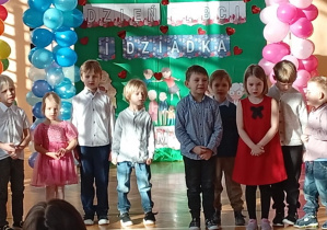 Dzieci stojące na sali gimnastycznej przed dekoracją z napisem Dzień Babci i Dziadka.