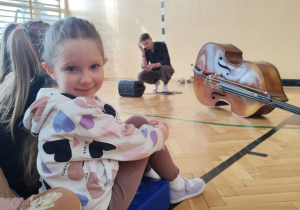 Uśmiechnięta dziewczynka siedząca na sali gimnastycznej podczas koncertu muzycznego.