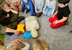 Grupa dzieci siedzących na dywanie przyglądająca się fantomowi podczas resuscytacji.