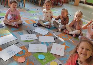 Grupka dzieci siedzących w kole na dywanie przy obrazkach i kartkach z napisami w języku angielskim
