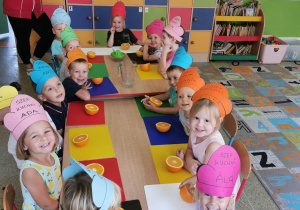Grupka dzieci wraz z panią siedząca przy stole przygotowuje sok pomarańczowy