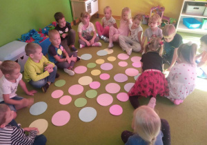 Grupa dzieci siedzących na dywanie patrzących na porozkładane kolorowe papierowe kropki