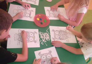 Kilkoro dzieci siedzących przy stole i stemplujących obrazem za pomocą farb oraz patyczków