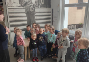 Grupka dzieci stojących przy ścianie słuchających pani bibliotekarki
