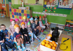 Dzieci z grupy biedronek siedzące na krzesełkach przed stolikiem na którym leżą pomarańcze,marchewki oraz sokowirówka.