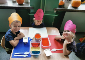 Grupka dzieci siedzących przy stoliku na którym leżą kolorowe galaretki w pojemniczkach.