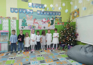 Grupa dzieci stojąca przy choince na tle dekoracji świątecznej.