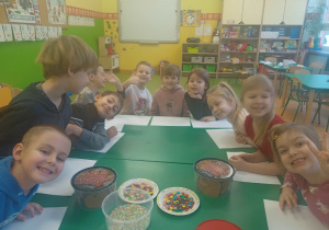 Grupa uśmiechniętych dzieci, siedzących przy stoliku, dekorują ciastka. Na stoliku leżą ciastka i kolorowe posypki.