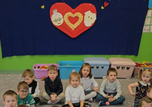 Grupa dzieci siedząca w kształcie serca na dywanie.