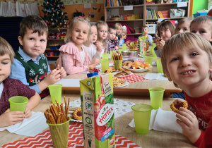 W sali przedszkolnej dzieci siedzą przy zastawiony słodyczami i napojami stole