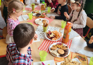 W sali przedszkolnej dzieci siedzą przy zastawiony słodyczami i napojami stole
