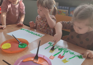 Dzieci wykonują prace plastyczna , trzymają w ręku widelec na stoliku talerzyki z kolorowymi farbami.